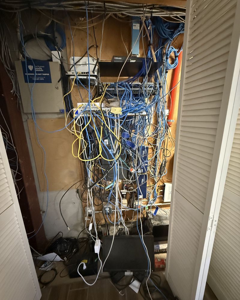 Network closet before remediation: unlabeled cable draped in loops across the whole closet, equipment buried behind it, gear stacked on the floor